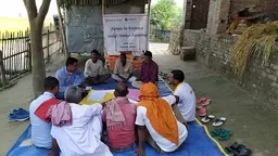 A group of girls sitting in a circle on a mat, having a discussion. In the background, a poster reads "Partners for Resilience through Strategic Partnership".