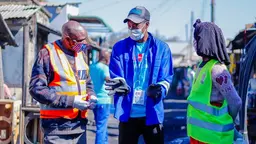 Three people stand talking in a street. A man, a woman and, in the middle, a young man wearing a face mask.
