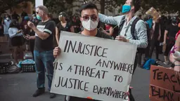 A masked protester holds up a sign saying "Injustice anywhere is a threat to justice everywhere"