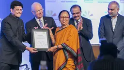 Prema Gopalan stands centre stage in an orange sari. Two men present her with a certificate; two others stand behind and applaud.