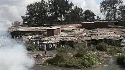 Closely built shacks. In the background, trees. In the foreground, a river and, on the other side of it, smoke.