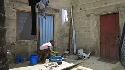 A woman sits in the courtyard of a building, washing clothes in a small bowl.