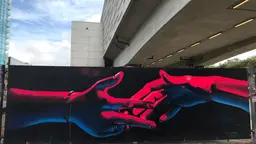 A bleak urban landscape. In the foreground, a painting of two hands reaching towards each other, painted in red and blue on a black background.