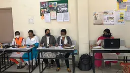 Five women, wearing masks, sit at desks in front of computers and mobile phones