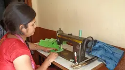 A young woman sits working at a sewing machine in a corner of a room.