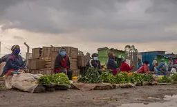 Women sitting next to vegetables wear masks and keep social distancing.
