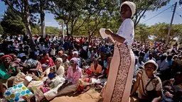 Woman stands in front of a large number of woman and children sat on the floor outside.