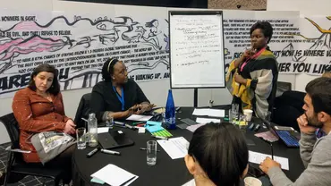 Men and women sit around a table listening to a woman talk next to a white flipboard. behind them is a giant illustration related to climate change.
