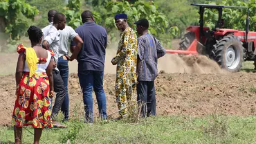 A group of five men and women gather and talk on a field, while a tractor ploughs the field in the background.