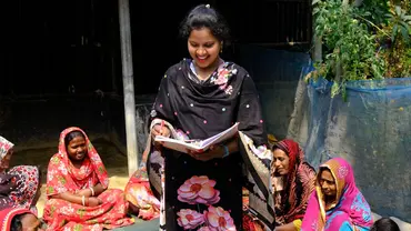 Group of women wearing tradition Bangla dress gathered in a circle to discuss. One of the women is standing up and reading from a notebook,smiling.