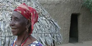 Old woman in profile in front of thatched hut and mud house. Niger. 