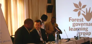Two men sat behind a desk chat to one another, with the Forest Governance Learning Group logo in the background