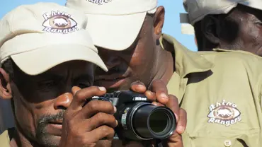 A man peers through the lens of a camera, while receiving advice from his colleague, another ranger
