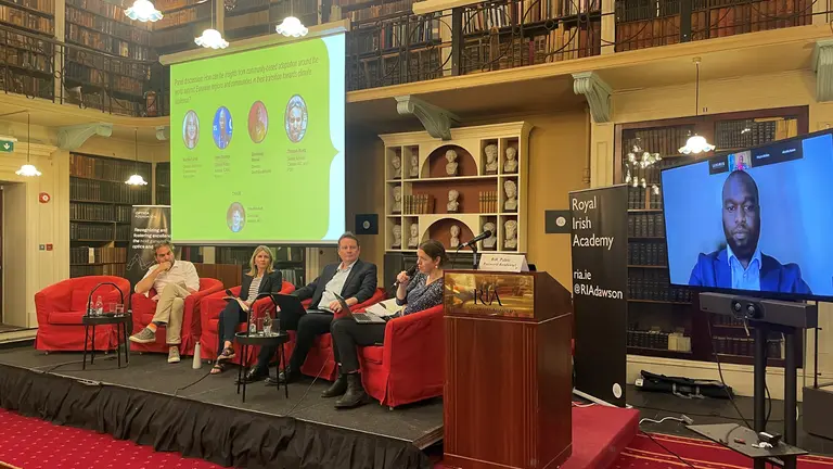 A seated panel of four people listen to a speech given by a remote colleague on a television screen positioned to their left.