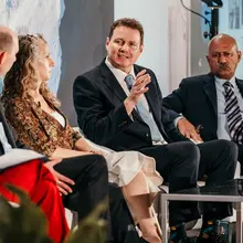 A suited man seated in a panel gesticulates as he makes his point while speaking.