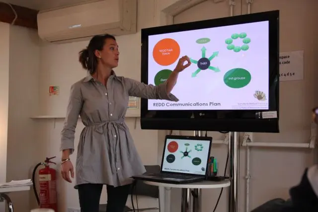 Woman stands at the front of a room pointing to a presentation on a screen behind her.