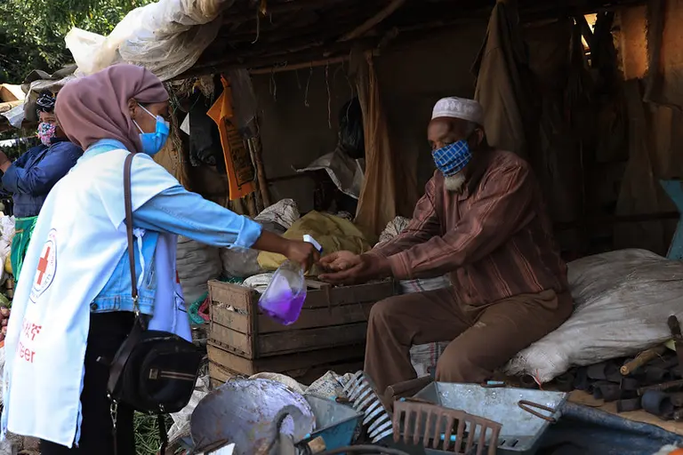 Young woman wearing a red cross vest sprays an old man hands. Both wear masks. 