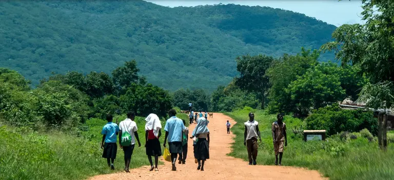 Children are pictured from the back walking along a dusty road, with others walking the other way and mountains in the background.