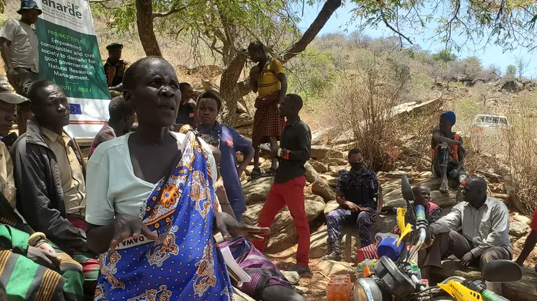 A woman stands to speak at a community meeting, surrounded by other people.