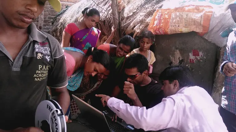A group of people gather around a man seated with a laptop computer and look and point at the screen