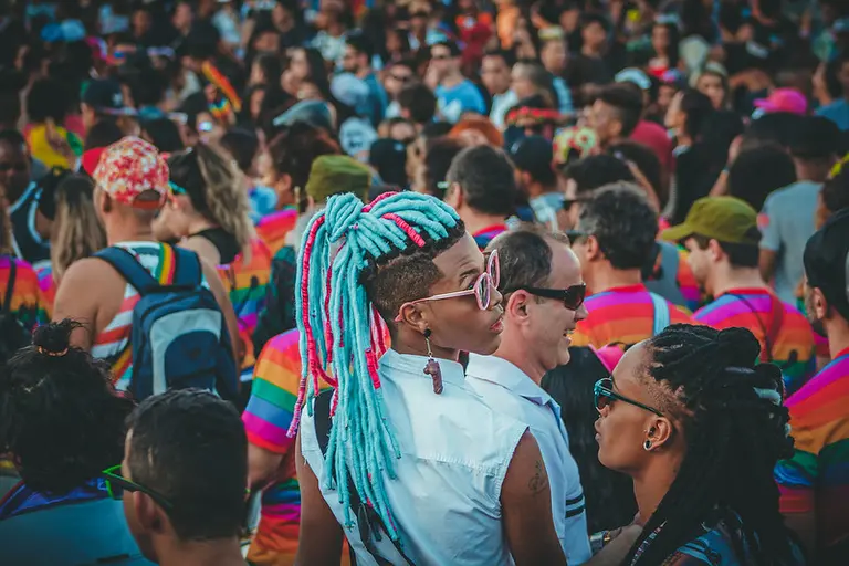 Young man with blue and pink dreadlocks surrounded by a crowd wearing pride colours