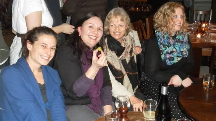 Woman sits among friends and smiles while posing for a photo, with drinks on the table in front of her.