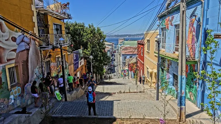 Street with colourful buildings and people standing in the shade. A body of water can be seen in the background.