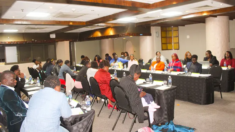 A group of people sat at tables in conversation with one another and listening to a presentation. 