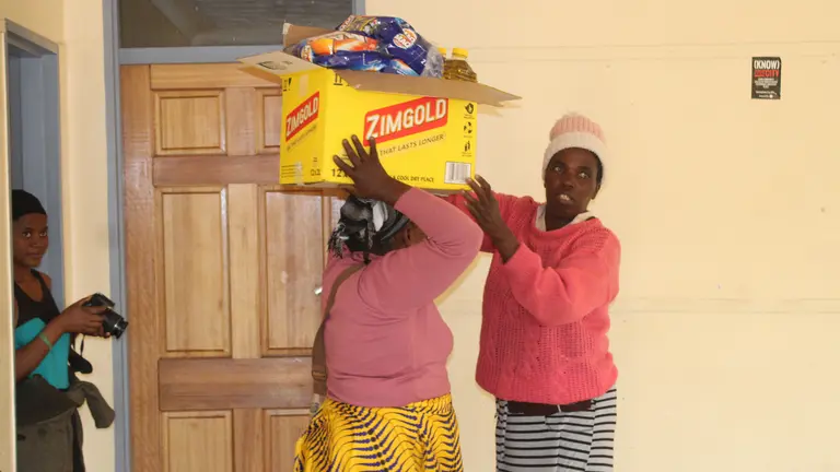 A woman helps another woman balance a box overflowing with groceries on her head, while a third woman watches from a doorway.