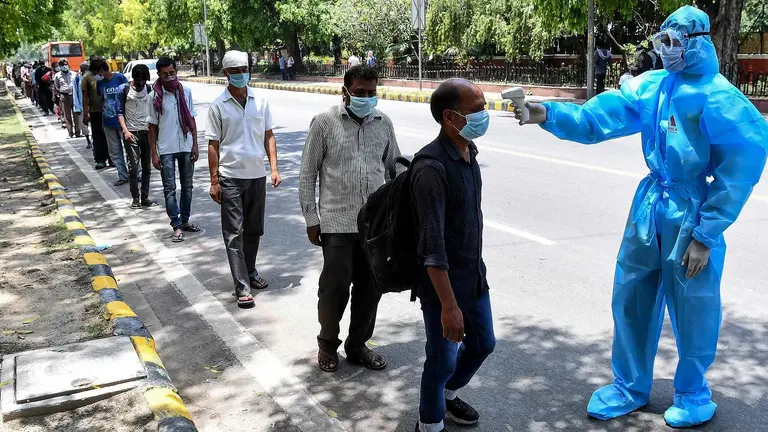 A line of people wearing masks, spaced apart, wait for a man dressed in full protective clothing pointing a temperature gun to test for COVID-19.