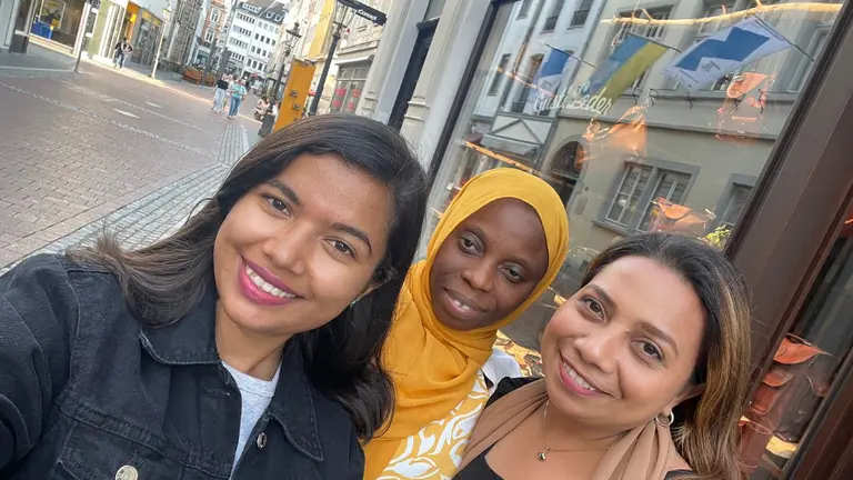 Three women smile as they pose for a selfie outdoors on a street.