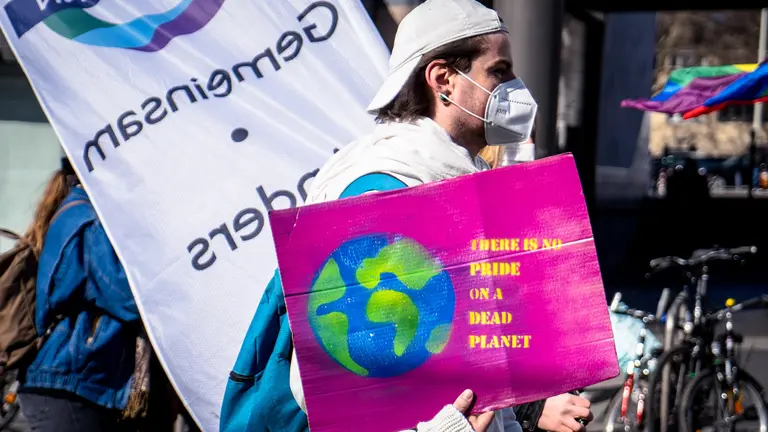Man wearing a mask on a protest march carries a sign, with a pink background, saying: "There is no pride on a dead planet".