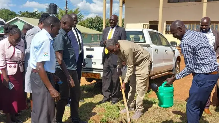 A group of people stand around a man with a spade planting a tree, with buildings in the background.