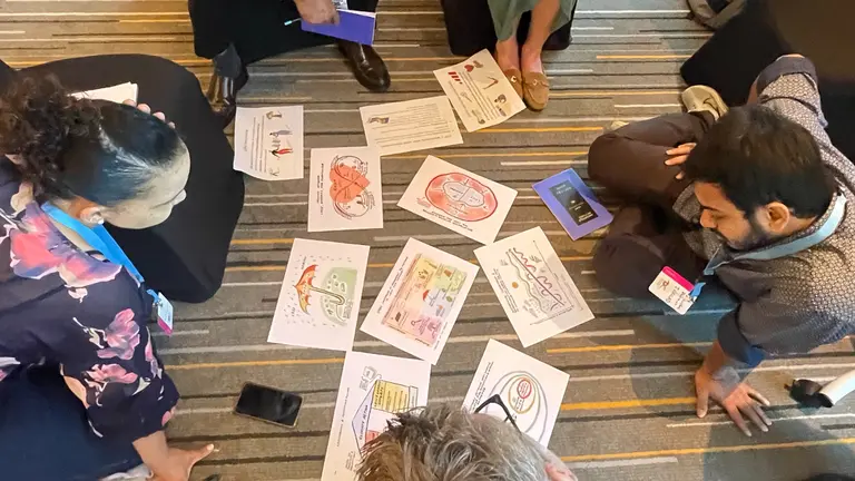 People sit on the floor around a series of printed cartoons and illustrations in the centre.