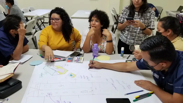 A group of individuals gathered around a table, talking, writing and drawing on a whtite piece of paper.eas.