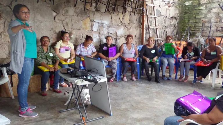 A woman stands and gives a speech in front of other women seated in a circle around her.
