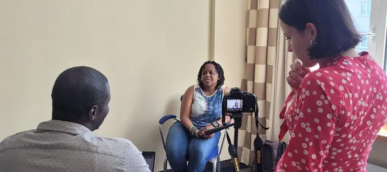 A woman seated in a chair faces a video camera, with a man and woman behind the camera looking quizzical. 