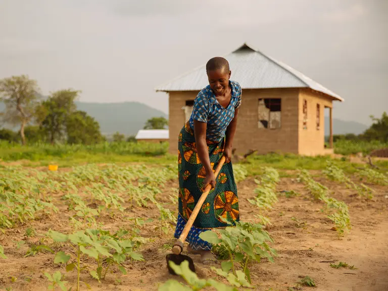 A woman farmer tends her crops with a hoe in front of a small house.