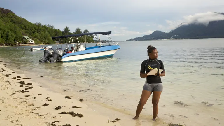 A young woman wearing shorts, standing on a beach in the Seychelles. There is a boat behind her.