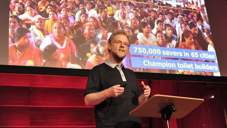 A bearded man stands at a podium with a screen behind him.