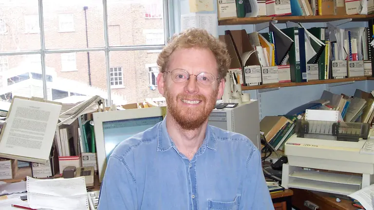 A man smiles for a photo while surrounded by a messy desk and multiple books and reports.