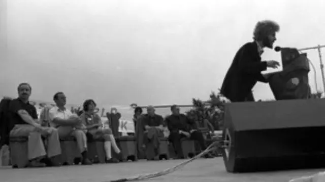 A black and white photo showing a man giving a speech at a podium with people seated behind him. 