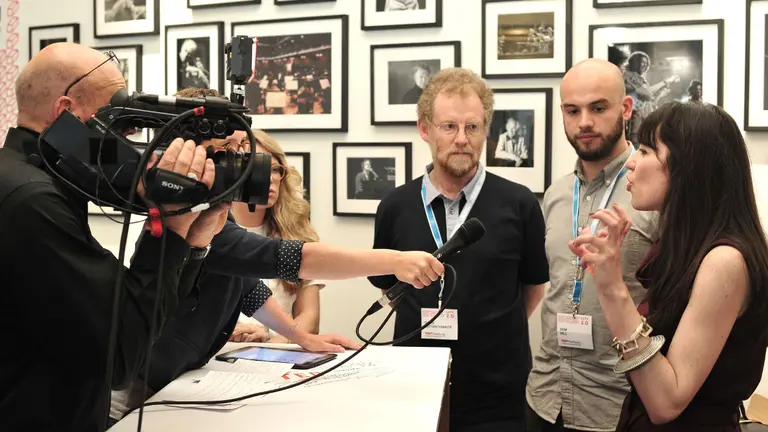 A group of men and women standing around a table are interviewed by a man with a camera and microphone.