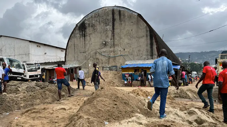 Men standing among large piles of sand. Concrete sheds behind them.
