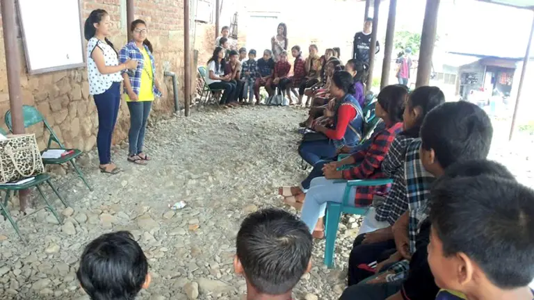 Two people stand in an outside area, surrounded by seated youngsters in a semi-circle.