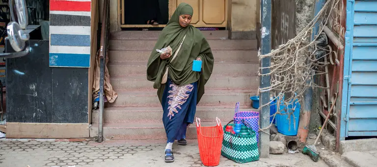 A woman in a headscarf stands outside a building, with two large bags at her feet.
