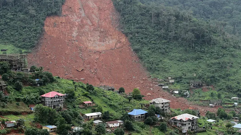 A huge landslide on the side of a hill, with small houses just beneath it.