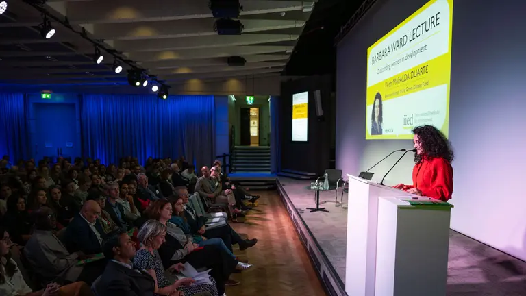 A woman stands at a podium in the foreground, while a large seated audience listens.  