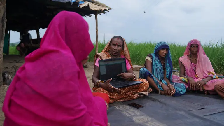 Women sitting down in a circle. One of them is holding a laptop and talking.