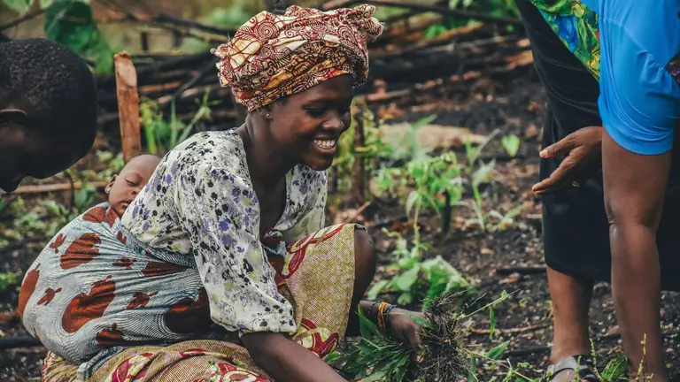 A woman picks out grass from a garden with baby sleeping on her back.